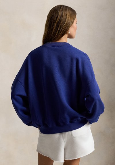 Φαρδιά μπλε σκούρα μπλούζα με rib λαιμόκοψη και μανσέτες, με χαλαρές ραφές στους ώμους και υφή στο ύφασμα, σε συνδυασμό με λευκά σορτς.