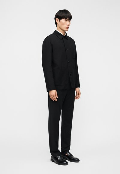 Μαύρο σακάκι με μεγάλα κουμπιά, επίπεδη γιακά και δύο μπροστινές τσέπες, σετ με ταιριαστό παντελόνι και μαύρα παπούτσια, παρουσιασμένο σε μινιμαλιστικό στυλ.