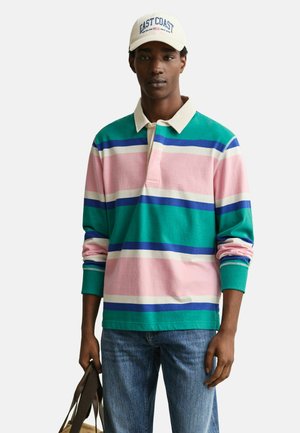 Young man wearing a striped long-sleeve rugby shirt in green, pink, blue, white, jeans, white cap reading "East Coast," holding a beige bag.