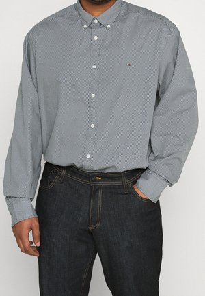 Homme portant une chemise à carreaux noir et blanc boutonnée, rentrée dans un jean bleu foncé, avec une main dans la poche.