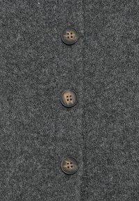 Šedá vlněná tkanina s třemi tmavě hnědými knoflíky, z nichž každý má dvoupodlažní design, na texturovaném pozadí.