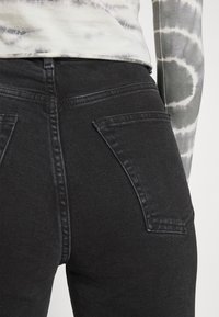 Vaqueros de denim negro con un diseño ajustado, que presentan bolsillos traseros y costuras en contraste. La textura parece suave con un ligero estiramiento.