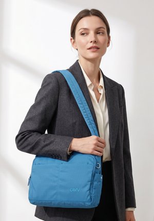 Donna con blazer scuro e camicetta bianca che porta una borsa a tracolla blu con il logo "GMV", in piedi davanti a uno sfondo chiaro e uniforme.