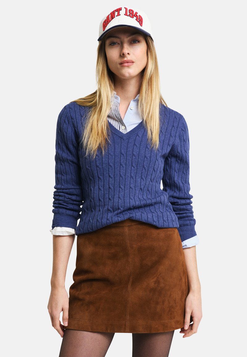 Marinefarbener Cable-Knit-Pullover über einem hellblauen Hemd mit Kragen, kombiniert mit einem braunen Wildleder-Minirock und einer weißen Mütze mit rotem Text.