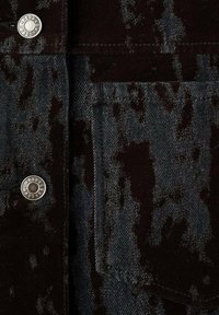 Tessuto in denim nero e scuro con un motivo variopinto, caratterizzato da bottoni in metallo argentato e un dettaglio di tasca applicata. Superficie testurizzata.