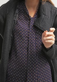 Persona che indossa una blusa a motivi blu navy e un cappotto nero con texture, mentre regola il cappotto con la mano destra vicino al colletto.