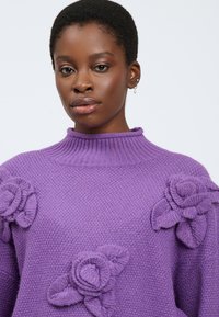Junge Frau mit kurzen Haaren, die einen lila Strickpullover trägt, der mit strukturierten Blumenmustern an den Schultern und der Brust verziert ist.