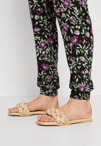 Sandalias planas trenzadas beige con textura tejida y suela lisa de color marrón claro. Combinadas con pantalones negros con un patrón floral que presenta acentos en púrpura y verde.