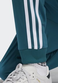 Los pantalones atléticos color verde azulado tienen rayas laterales blancas, puños acanalados y una textura de tela suave. Se combinan con zapatillas Samba blancas y calcetines con estampado.