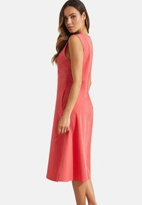 Robe sans manches corail avec un corsage ajusté, une jupe évasée et des poches latérales. Le tissu semble lisse avec un léger éclat.