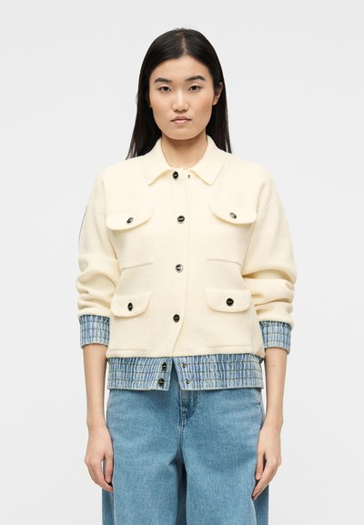 Femme portant une veste crème avec des boutons noirs sur une chemise à carreaux bleue, assortie à un jean ample bleu clair, debout devant un fond blanc.