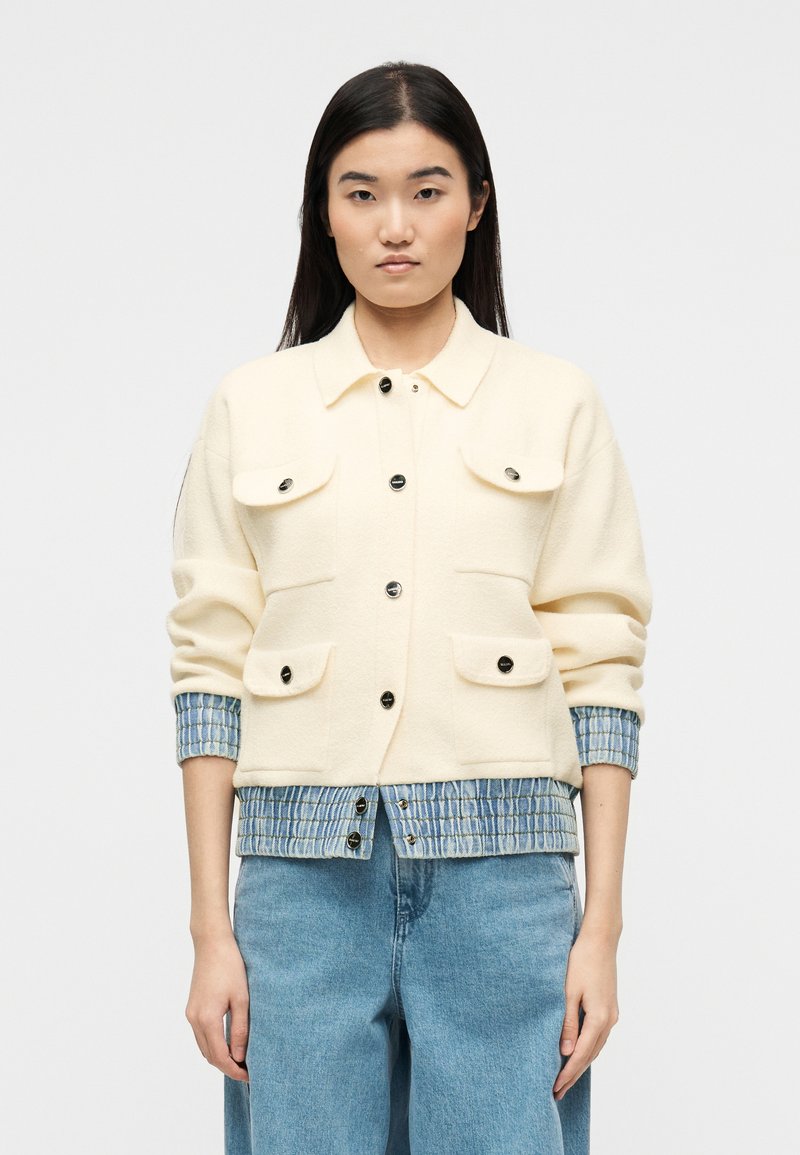 Γυναίκα που φοράει κρεμ μπουφάν με μαύρα κουμπιά πάνω από μπλε πουκάμισο με καρό μοτίβο, συνδυασμένο με ανοιχτόχρωμα μπλε φαρδιά τζιν, όρθια μπροστά σε λευκό φόντο.