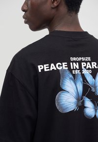 Person trägt ein schwarzes T-Shirt mit weißem Text "DROPSIZE PEACE IN PARADISE EST. 2020" und drei großen blauen Schmetterlingen auf dem Rücken.