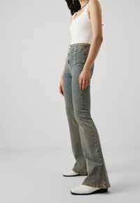 Ljusblå högmidjade jeans med utsvängda ben, featuring två framknappar och subtila sömnadsdetaljer; ihop med vita ankelstövlar.