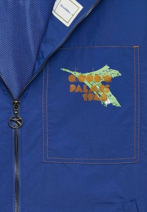 Blå jakke med lynlås, mesh-foring og en broderet design med "Palmes 1948" i orange og grøn på brystlommen.