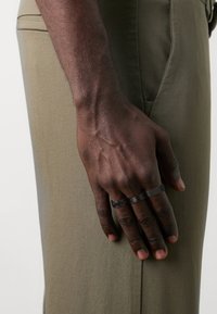 Svart hånd hvilende på khaki bukser, med tre ringer: en med mesh-design, en glatt, og en med kjede-lignende mønster.