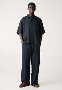 Camicia a maniche corte in lino blu scuro con bottoni frontali e tasca applicata, abbinata a pantaloni larghi dello stesso colore e sandali scivolati marroni.