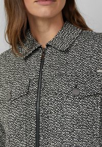 Kobieta w czarno-białej kurtce z teksturowanym materiałem, zapinanej na zamek z kołnierzem i dwiema kieszeniami na guziki na piersi.