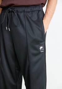 Person iført sorte Nike Air sweatpants med elastisk talje og snøre, hånd i sidelommen, mod en ensfarvet lys baggrund.