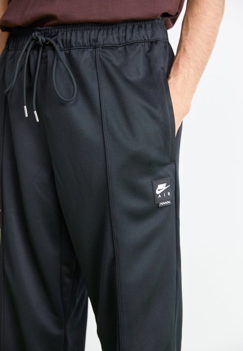 Persona che indossa pantaloni neri Nike Air con cintura elastica e cordino, mano nella tasca laterale, su uno sfondo chiaro e semplice.