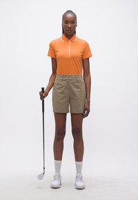 Femme en polo orange, short beige et chaussures de golf blanches tenant un club de golf sur un fond blanc uni.