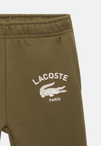 Tecido verde-oliva com cintura elástica, apresentando o logótipo branco da Lacoste e o símbolo do crocodilo acima da palavra Paris.