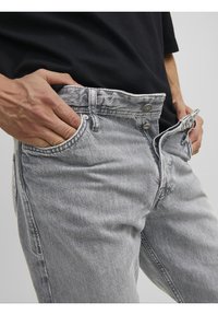 Jeans de mezclilla gris con una textura desgastada, estilo de cinco bolsillos y cierre de cremallera. Presentan herrajes metálicos y un acabado ligeramente desteñido.