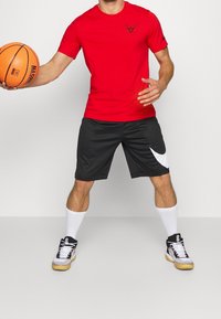 Mann in rotem Chicago Bulls Shirt, schwarzen Nike Shorts, weißen Socken und Turnschuhen, der einen Basketball hält, während er in athletischer Pose steht.