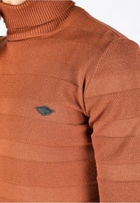 Pull en maille marron texturé à col roulé avec rayures horizontales et petit logo noir brodé sur le côté gauche de la poitrine.