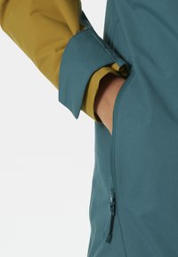 Wasserabweisende Jacke mit einem blau-grünen Körper und gelb-grünen Ärmeln, seitlichem Reißverschlussfach und kontrastierenden Details an den Bündchen.