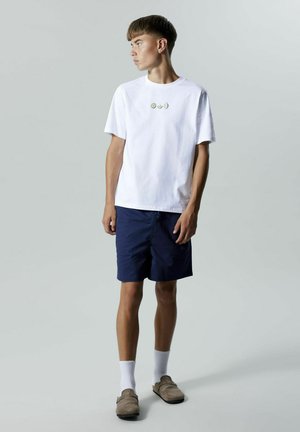 T-shirt bianco in cotone con tre piccole decorazioni di frutta, abbinato a pantaloni corti blu navy. La calzatura è composta da scarpe slip-on beige chiaro con una texture morbida.