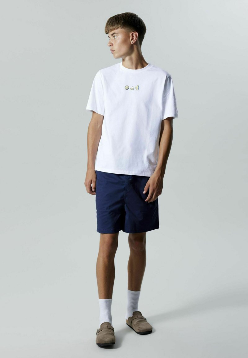 Hvid bomulds t-shirt med tre små frugtmotiver, sammen med marineblå shorts. Fodtøjet er letbrune slip-on sko med en blød tekstur.