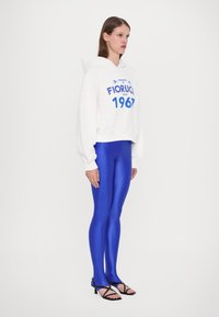 Hvit, kort hoodie med "Fiorucci 1967" trykk, kombinert med glansfulle blå leggings og svarte sandaler med stropper. Jevn stoffkvalitet og avslappet passform.
