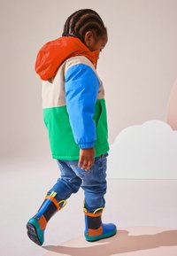 Chaqueta impermeable colorida con capucha naranja, secciones en verde, azul y beige; combinada con jeans azules y botas de goma multicolores.