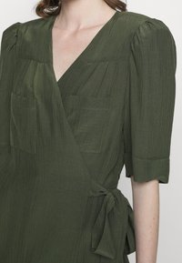 Olive green wrap dress made of textured fabric, featuring short puffed sleeves, two chest pockets, and a tied waist detail.