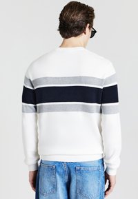 Maglione bianco con strisce orizzontali in blu scuro, grigio chiaro e bianco. Il design include un colletto e dei polsini a costine, abbinati a pantaloni in denim blu.