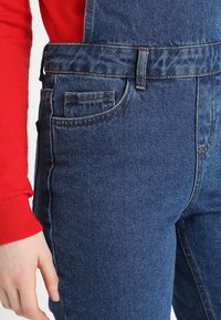 Salopette in denim blu con tasca frontale, spalline spesse e design aderente. Il tessuto presenta una leggera trama e dettagli di cuciture sottili.