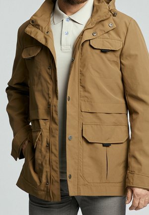 Homme portant une veste à capuche couleur sable avec plusieurs poches à rabat à l'avant, par-dessus un polo beige clair et un pantalon gris à motifs.