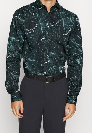 Chemise noire à manches longues avec un motif marbré vert et blanc, dotée d'un col pointu et d'une fermeture à boutons, associée à un pantalon sombre et une ceinture noire.