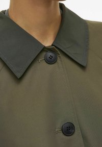 Kadar u bližem planu prikazuje osobu koja nosi maslinasto zeleni kaput s tamnozelenim špicastim ovratnikom i dva crna dugmeta zakopčana sprijeda.