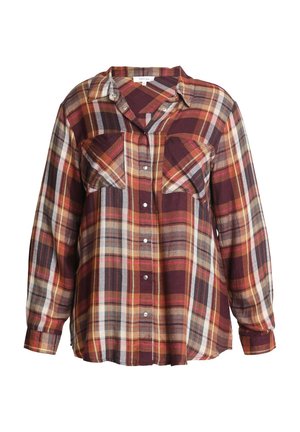 Vrouw draagt een geruite blouse met knopen en lange mouwen in bordeaux-, oranje- en gele tinten, met één hand in de zak van een blauwe spijkerbroek.