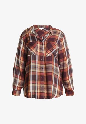 Femme portant une chemise à carreaux boutonnée à manches longues dans des tons bordeaux, orange et jaune, une main dans la poche d'un jean bleu.