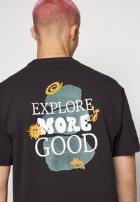 Personne aux cheveux courts roses portant un t-shirt noir avec le texte "Explore More Good" et de petites créatures de dessin animé jaunes dans le dos.