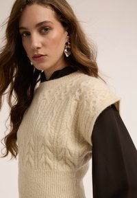 Kremowy bezrękawnik z dzianiny z warkoczowym wzorem, zestawiony z czarną, długorękawową bluzką. Modelka ma fale na włosach i duże kolczyki.