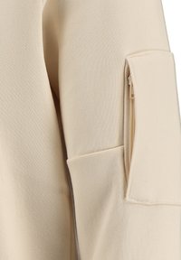 Top beige de manga larga con un tejido texturizado, bolsillo con cremallera en la manga y un ligero detalle en los puños.