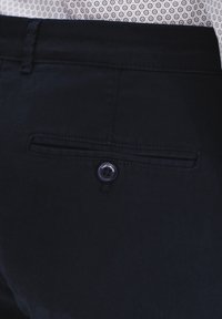 Ciemnogranatowe spodnie z tylną kieszenią z zapięciem na guzik. Tkanina jest gładka, z delikatnym połyskiem i minimalnie widocznymi szwami.