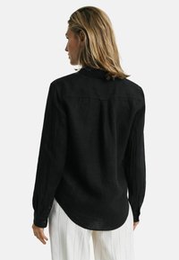 Svart långärmad skjorta i texturerad tyg, med en knäppningkrage, bakre yoke-seam och manschetter med knappar, bärs med vita randiga byxor.