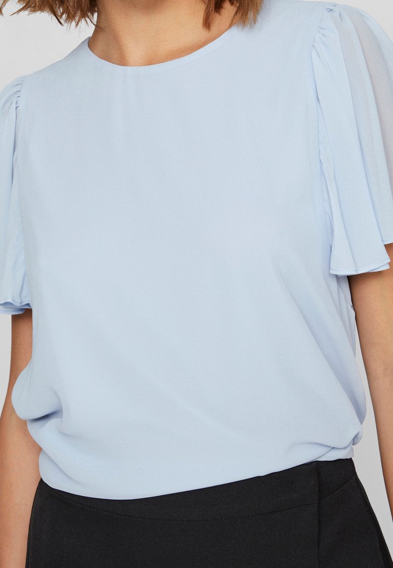 Blouse bleu clair à manches courtes évasées, portée rentrée dans un pantalon noir taille haute sur une personne aux cheveux mi-longs.