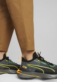 Person, der braune Hose und schwarze, grüne, gelbe Puma Sneaker trägt, steht vor einem hellen Hintergrund.