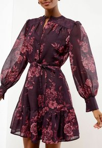 Donna che indossa un vestito floreale viola scuro con maniche lunghe a sbuffo, frontale con bottoni, cintura in vita e orlo arriccicato.